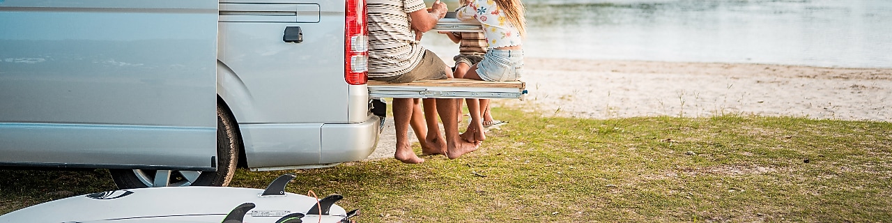 A young Caucasian family enjoys a casual meal on their minivan’s foldable dining table by a calm seaside, surrounded by nature and surfboards.