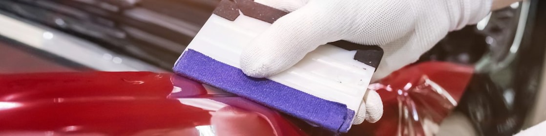 A gloved hand smoothing paint protection film on a red car.