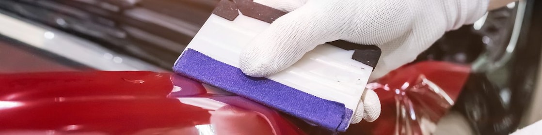 A gloved hand smoothing paint protection film on a red car.