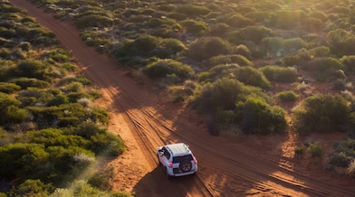 A truck navigates a rugged dirt road, surrounded by the vast expanse of a sunlit desert landscape.