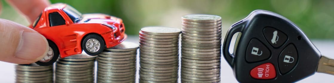 A hand places a toy car is over a stack of coins with a car key.