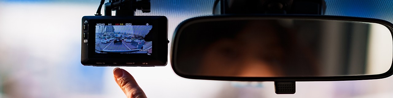 A hand points at a dash cam display located on a front windscreen to the left of the rear-view mirror.