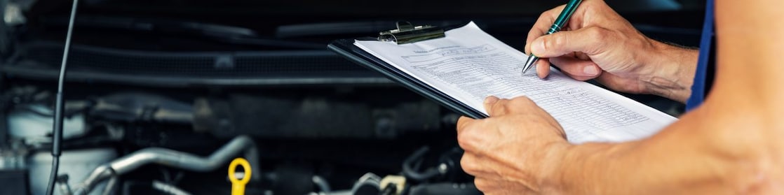 Close up of mechanic checking off notes on a clipboard in front of an open car bonnet.