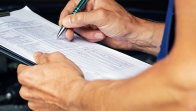 Close up of mechanic checking off notes on a clipboard in front of an open car bonnet.