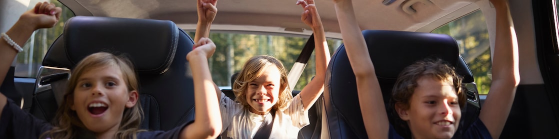 3 kids in the back of the car with seatbelts on and with their hands up looking excited