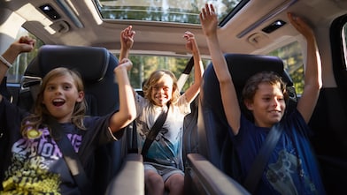 3 kids in the back of the car with seatbelts on and with their hands up looking excited