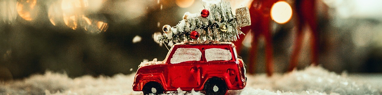 A miniature Christmas scene showing snow falling on a metallic red toy car with a Christmas tree on top.