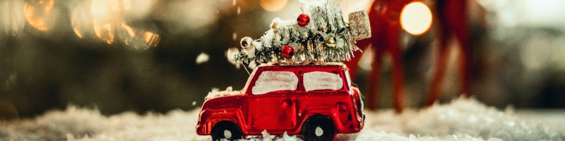 A miniature Christmas scene showing snow falling on a metallic red toy car with a Christmas tree on top.