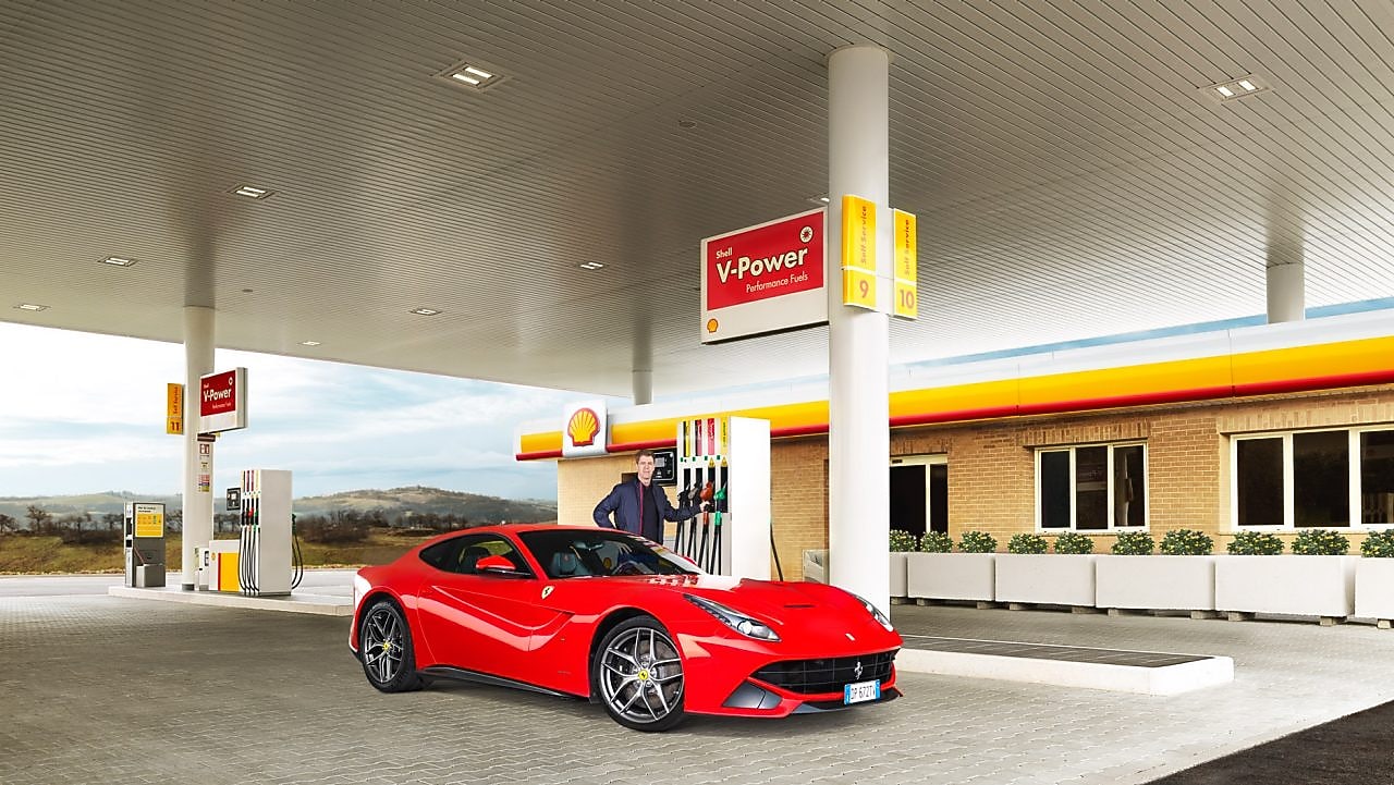 A red Ferrari sitting on a Shell station forecourt with a man leaning on a petrol pump