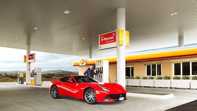 A red Ferrari sitting on a Shell station forecourt with a man leaning on a petrol pump
