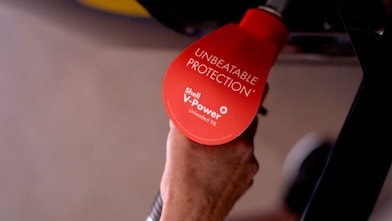 Close up of a fuel nozzle refuelling a car with Shell V-Power 98. The fuel nozzle decal says: Unbeatable Protection.