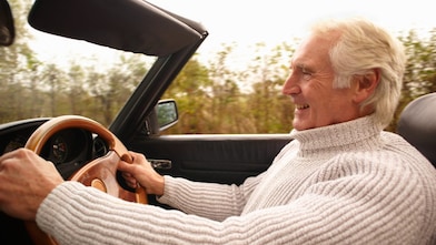 A man drives an open-top car along a tree-lined road