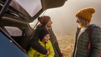 A young family of three stand by the boot of their 4WD wearing beanies and winter clothing, parked by a foggy field.