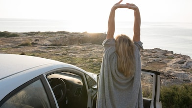 A woman stands by her car, stretching her arms in the air
