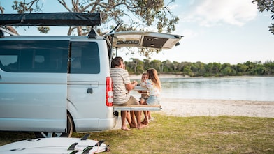 A young Caucasian family enjoys a casual meal on their minivan’s foldable dining table by a calm seaside, surrounded by nature and surfboards.