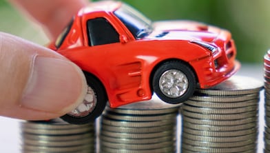 A hand places a toy car is over a stack of coins with a car key.